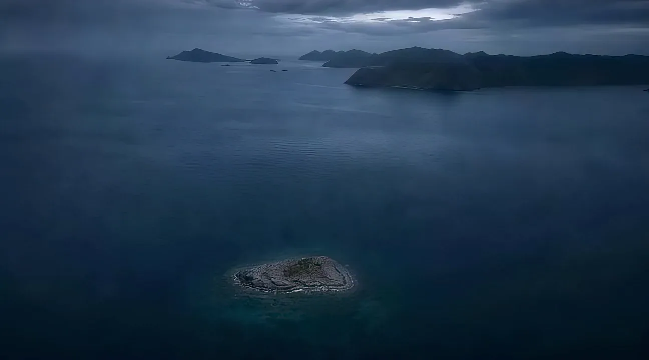 A single very small island far out in open water, with a larger island chain on the horizon.
