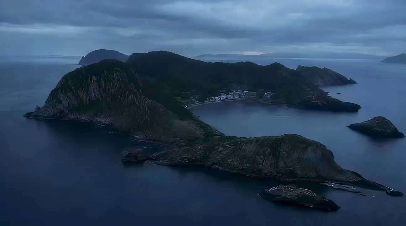A large headland island curving around a sheltered bay, with a small settlement gathered at the waterline.
